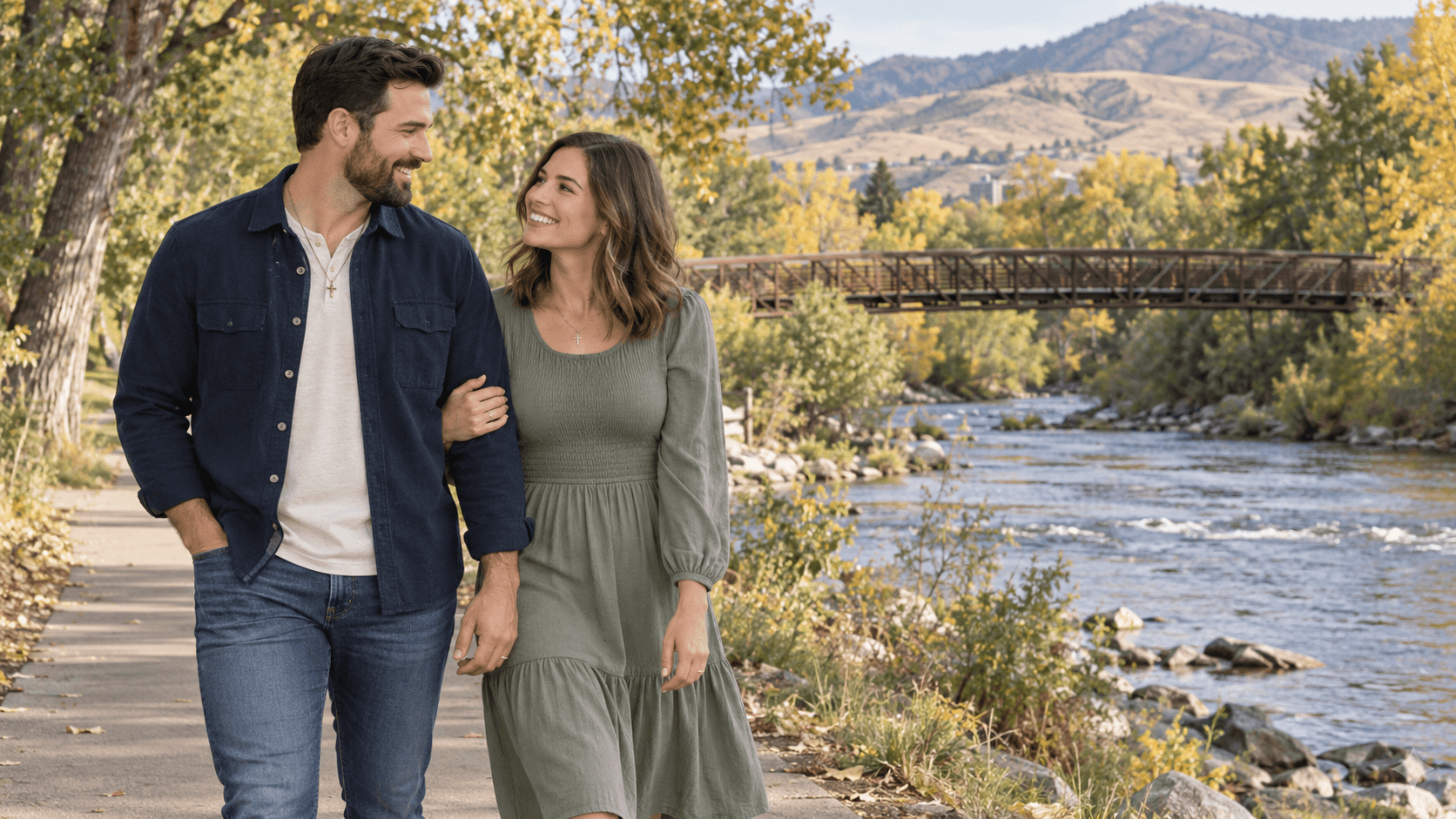 Couple wearing cross necklaces walking beside a river and autumn trees in a Boise Greenbelt-inspired scene.